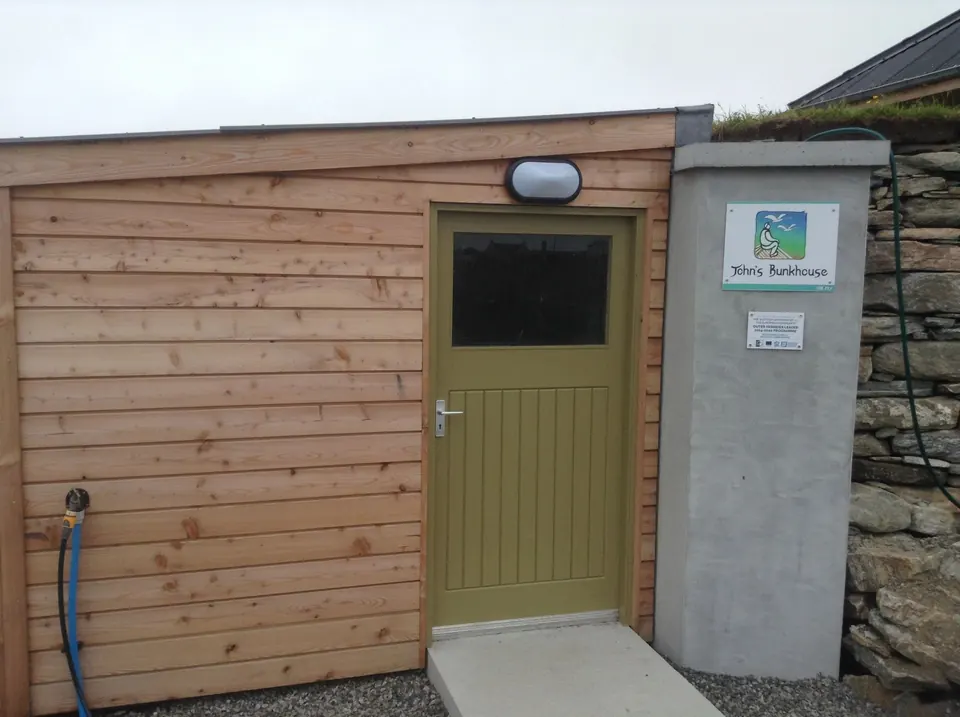 The timber-clad bike shed with a green door, beside the stone wall of the bunkhouse