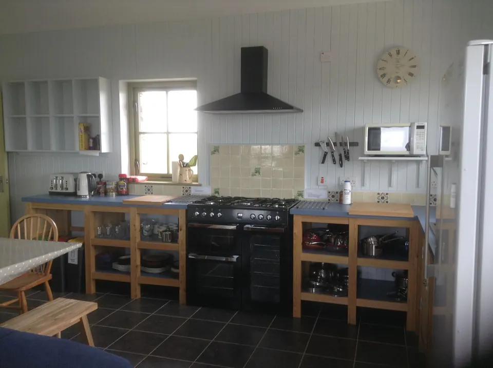 The kitchen: a range cooker under an extractor hood, open timber shelving stocked with pans and crockery, and a wall clock