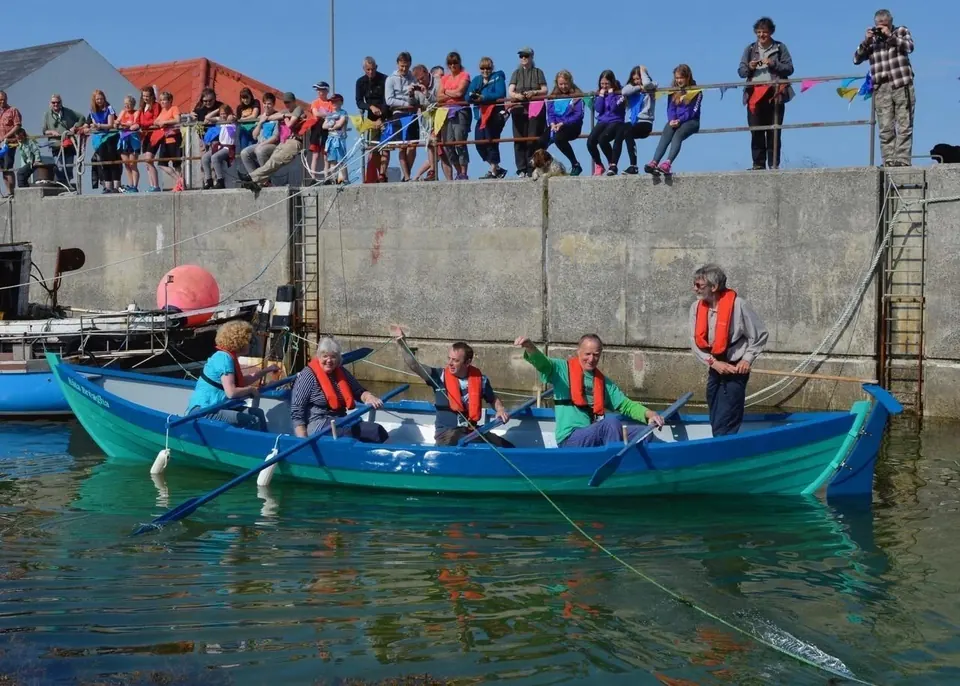 A crew in life jackets rowing a blue skiff out of the harbour while a crowd watches from the quay